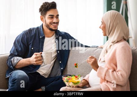Schwanger Islamische Frau Essen Gemüsesalat, Während Sie Sich Auf Der Couch Ausruhen Mit Ehemann Stockfoto