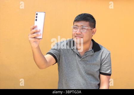 Junger, dunkelhäutiger Latino-Mann mit Brille benutzt sein Handy, um online zu gehen, Anrufe zu tätigen, Nachrichten zu senden, einzukaufen und Termine zu vereinbaren Stockfoto