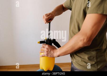 Hausmeister männlich pumpt gelbe Wasserspritzflasche beim Halten auf blauem Sofa, Vorbereitung zur Reinigung oder Desinfektion von Polstern in der häuslichen Umgebung. Stockfoto