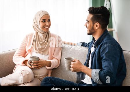 Fröhliches, Schwanger-Muslimisches Paar, Das Sich Zu Hause Ausruhte, Kaffee Trinkt Und Sich Unterhielt Stockfoto