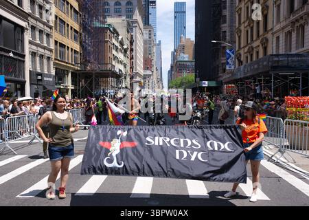 Eine Straßenszene bei der NYC Pride Parade, die zwei Frauen zeigt, die ein Banner für „Sirens MC NYC“ halten, einen Mann in Badehosen, Zuschauern und Motorrädern. Ta Stockfoto
