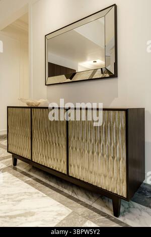 Ein stilvolles Sideboard aus Holz mit strukturierten goldenen Türen und einem abgeschrägten Spiegel darüber. Elegante Möbel auf Marmorboden in einem luxuriösen Eingangsbereich. Stockfoto