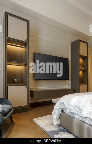 Modernes Schlafzimmer mit einem großen Fernseher an einer strukturierten Wand, flankiert von individuell eingerichteten Regalen und Schränken mit integrierter Beleuchtung. Stockfoto