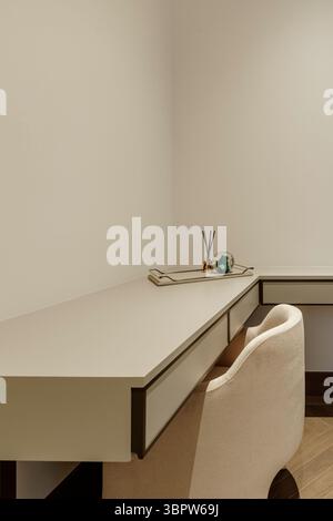 Nahaufnahme eines modernen, minimalistischen grauen Schreibtisches in einer Ecke. Ein dekoratives Tablett bietet Platz für einen Reed-Diffusor und einen Briefbeschwerer aus grünem Glas. Stockfoto
