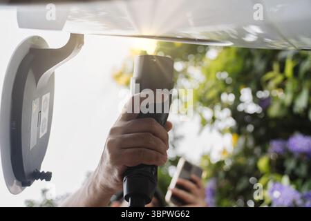 Nahaufnahme Mann, der das Ladegerät für Elektrofahrzeuge anschließt, während er das Telefon hält, um das intelligente Aufladen zu aktivieren Stockfoto