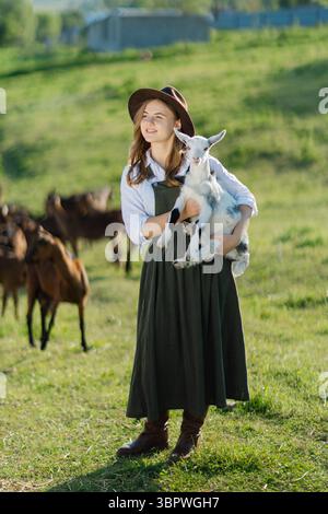 Eine junge Bäuerin mit Hut, eine Ziege in den Armen, auf einem Feld mit anderen Ziegen Stockfoto