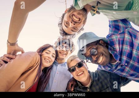 Eine vielfältige und glückliche multiethnische Gruppe junger Freunde, die sich in einem Kreis versammelten, lachten und lächeln, während sie aus einem niedrigen Winkel auf die Kamera blicken Stockfoto