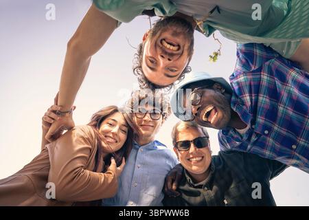 Eine vielfältige und glückliche multiethnische Gruppe junger Freunde, die sich in einem Kreis versammelten, lachten und lächeln, während sie aus einem niedrigen Winkel auf die Kamera blicken Stockfoto