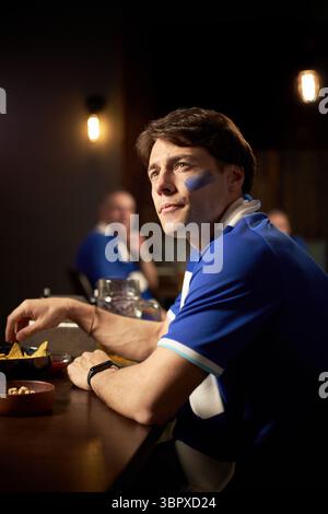 Kaukasischer junger Mann, der am Tisch sitzt und beim Fußballspiel zusieht, mit einem Gesicht in Mannschaftsfarben bemalt, Snack hält und mit einem anderen männlichen Fan im Hintergrund fokussiert aussieht Stockfoto
