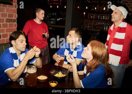 Gruppe von multiethnischen Fußballfans jungen und mittleren Alters, die Spiele in der Bar beobachten, Männer und Frauen sitzen und stehen um den Pub-Tisch mit Getränken und Snacks, konzentriert auf den Bildschirm Stockfoto