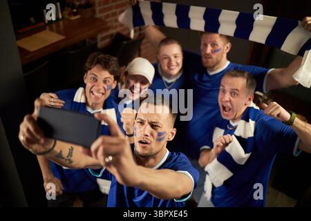 Gruppe multiethnischer Fußballfans, die gemeinsam Selfie feiern und machen, Männer und Frauen mit bemalten Gesichtern jubeln und lächeln, tragen Mannschaftstrikots und -Tücher Stockfoto