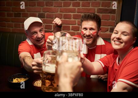 Eine Gruppe junger kaukasischer Fußballfans feiert gemeinsam, lächelt, toast und klirrt Bier, während man in der Bar das Fußballspiel ansieht Stockfoto