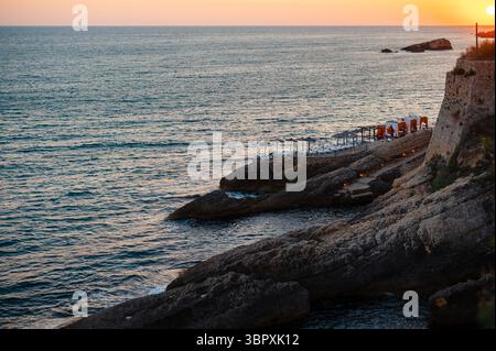 Eine ruhige Küstenlandschaft bei Sonnenuntergang mit felsigen Klippen und ruhigem Wasser. Am Ufer stehen Liegestühle und Sonnenschirme mit sanften Wellen Stockfoto