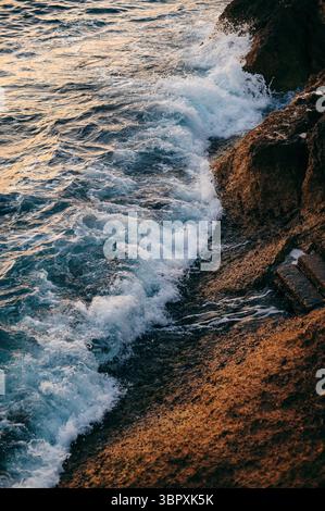 Wellen, die bei Sonnenuntergang gegen die felsige Küste krachen, schaffen eine dynamische und strukturierte Szene. Stockfoto