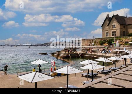 Lebhafte Sommerszene in einem Badebereich am Meer in Cascais, Portugal. Die Menschen entspannen und sonnen sich auf Steinterrassen, während andere im natürlichen po schwimmen Stockfoto