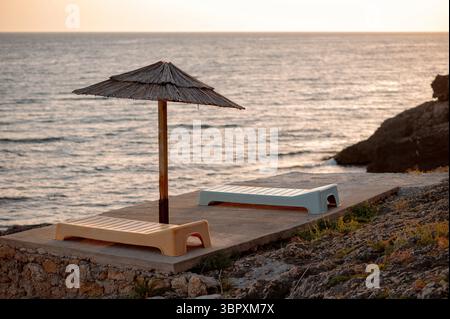 Zwei leere Liegestühle unter einem Strohschirm an einem felsigen Strand bei Sonnenuntergang. Stockfoto