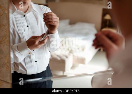 Nahaufnahme eines Bräutigams, der sich auf seinen Hochzeitstag vorbereitet. Der Mann berührt die Manschettenknöpfe auf seinem weißen Hemd mit der Reflexion im klassischen Hotelzimmer Stockfoto