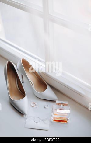 Nahaufnahme der Hochzeitsdetails. Bride Elegance Schuhe, Ringe, Parfum und Invation layflat auf weißer Fensterbank Stockfoto