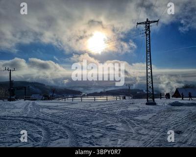 Ein großer Power-Pylon, der sich vor einem dramatischen sonnendurchfluteten Himmel befindet und einen starken Kontrast zwischen Technologie und Natur in einer verschneiten tschechischen Landschaft schafft. Stockfoto
