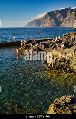 Menschen schwimmen in natürlichen Küstenbecken mit Los Gigantes Bergen und Klippen in der Ferne, Charco de Isla Cangrejo, Teneriffa, Kanarische Inseln, Spanien Stockfoto