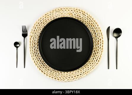 Draufsicht auf braune Korbtischmatte, schwarze Platte, schwarzes Besteck auf weißem Hintergrund. Rundes Strohfach, flache Ladefläche, Kopierraum. Stockfoto