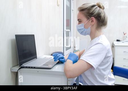 Zahnarzthelferin oder Krankenschwester, die in der Zahnarztpraxis mit medizinischen Handschuhen und Maske am Laptop arbeiten. Junger Arzt in weißer Uniform am Schreibtisch in der Klinik Stockfoto