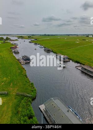 Eine ruhige Luftszene mit schwimmenden Häusern auf einem Fluss, umgeben von weiten grünen Wiesen und sich kreuzenden Wasserstraßen, mit niederländischen Fahnen, die sanft in der winken Stockfoto