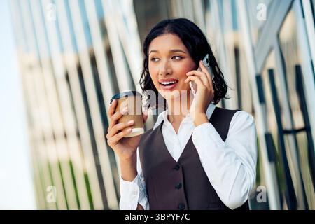 Professionelle Geschäftsfrau spricht am Telefon und hält Kaffee, was den modernen Erfolg in einer Unternehmensstadt repräsentiert Stockfoto