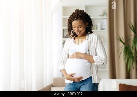 Schwarze schwangere Frau, die auf den Bauch blickt und lächelt, während sie in der Nähe des Fensters steht Stockfoto