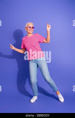 Modische Seniorin, die Freizeitmomente vor leuchtendem violettem vertikalen Hintergrund genießt und in lässiger, stilvoller Kleidung gekleidet ist Stockfoto