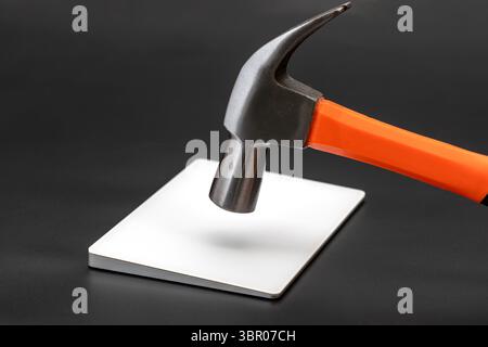 Ein Hammer mit orangefarbenem Griff befindet sich über der Metallplatte auf schwarzer Oberfläche, was auf einen drohenden Aufprall hindeutet. Die Szene vermittelt ein Gefühl der Vorfreude Stockfoto
