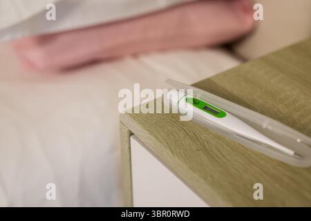 Digitalthermometer, offen auf hölzernem Nachttisch, Anzeige grüner Messwerte in der Nähe des rosafarbenen Kopfkissens. Gesundheit, Medizin, Diagnostik, Gesundheitswesen, modern, Stockfoto