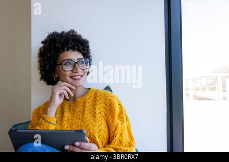 Afroamerikanerin mit Brille und Senfpullover, die auf dem Fenstersitz sitzt und ein Tablet hält. Gemütlich, modern, Lifestyle, Interieur, lässig, rev Stockfoto