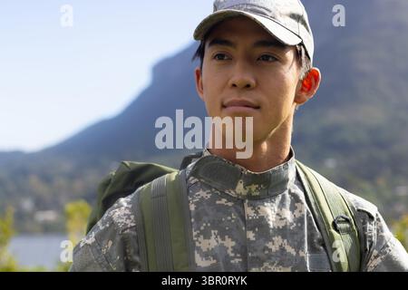 Asiatischer Soldat in Tarnmütze am See in der Nähe von Bergen mit grünem Rucksack, Kopierraum. Kadett, Abenteuer, Erkundung, Outdoor, Widerstandsfähigkeit Stockfoto