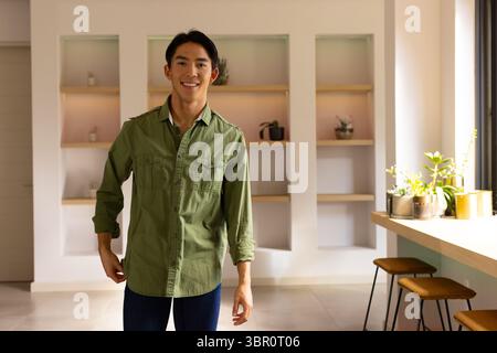 Asiatischer Mann lächelt, während er im Wohnbereich mit Wandregalen, Topfpflanzen und Barhockern steht. Modern, Lifestyle, minimalistisch, Interieur, Grün, entspannen Sie sich Stockfoto