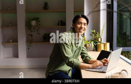 Asiatischer Mann, der am silbernen Laptop am Schreibtisch im Heimbüro mit Topfpflanzen und Kopfhörern arbeitet. Arbeitsbereich, Produktivität, Grün, modern, minimalistisch, Stockfoto