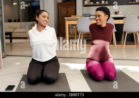 Verschiedene weibliche Freunde kniend auf Matten im Wohnzimmer mit Smartphone neben Matten in der Nähe des Sofas. Fitness, Wellness, Gesellschaft, lässig, modern, Leben Stockfoto