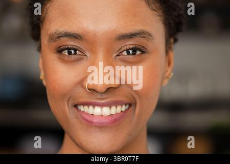 Afroamerikanerin lächelt und zeigt kurze lockige Haare, Nasenreif und Ohrstecker im Zimmer. Porträt, Schönheit, Schmuck, Schönheitsaufnahme, warme Töne, Stockfoto