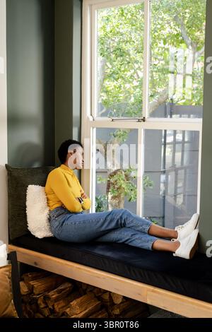 Frau mit Senfplatte, lehnt sich auf eine grüne Fensterbank mit Brennholz und blickt auf den Baum. Gemütlich, wohnlich, wohnlich, modern, rustikal, kontemplativ, s Stockfoto