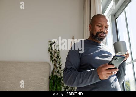 Afroamerikaner mittleren Alters, der zu Hause am Fenster steht, mit einer Pflanze, die Smartphone und Becher hält. Besinnlich, gemütlich, modern, Ruhe, Lifestyle Stockfoto
