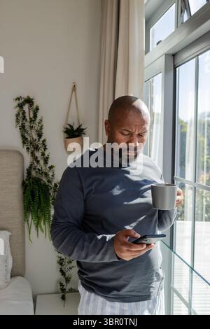 Afroamerikanischer Mann, der neben dem Bett am Fenster steht und einen grauen Becher hält und den Smartphone-Bildschirm scrollt. Modern, Entspannung, gemütlich, innen, Wellness Stockfoto