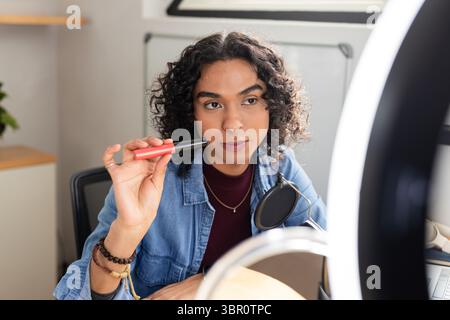Nicht binärer Erwachsener hält rote und schwarze Make-up-Röhre im Heimstudio unter Ringlicht mit Mikrofon. Kreativ, Technologie, modern, Empowerment, Innovation, ae Stockfoto