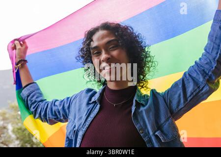 Nicht binäre Jeansjacke für Erwachsene, die die Arme im Park hochhebt und die mit Regenbogenstreifen gestreifte Stolz-Flagge hält. Vielfalt, Feier, Aktivismus, Jugendliche, Outdoor, vi Stockfoto
