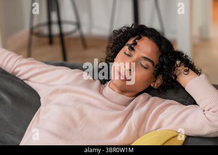 Nicht binäre Person, die sich auf einem grauen Sofa im Wohnzimmer mit senfgelben Kissen zurücklehnt. Modern, entspannend, minimalistisch, gemütlich, innen, Komfort, l Stockfoto
