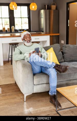 Ranghoher afroamerikanischer Mann im Sweatshirt, der Smartphone hält und auf dem Sofa neben der Frühstücksbar sitzt. Lifestyle, Entspannung, gemütlich, modern, häuslich, Co Stockfoto