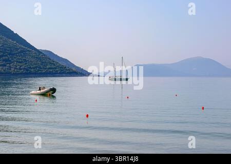 Ein Segelboot und ein Schlauchboot liegen im ruhigen, reflektierenden Wasser einer Bucht, mit trüben Bergen im Hintergrund unter weichem Himmel am Vasiliki Beach. Stockfoto