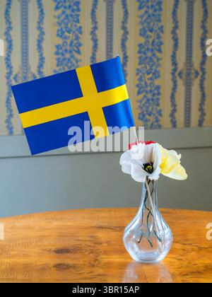 Eine kleine schwedische Flagge in einer Glasvase mit künstlichen Blumen auf einem Holztisch. Der Hintergrund ist mit einem gemusterten Hintergrund mit blauem A versehen Stockfoto