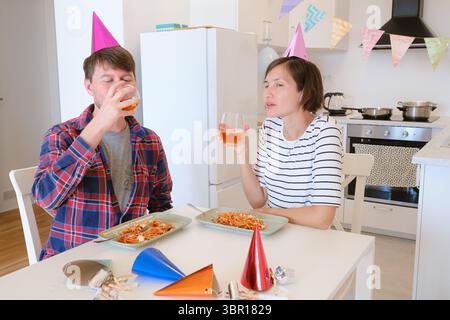 Das junge Paar feiert zu Hause gemütlich Geburtstag und genießt Kuchen und Lachen in warmer Atmosphäre. Die Familie isst Pasta und trinkt Wein zum Feiern Stockfoto