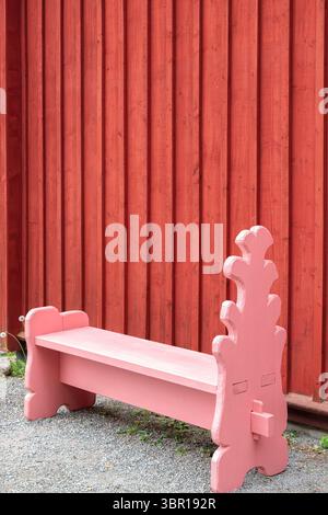Eine pinkfarbene Holzbank vor einer roten Holzwand, die eine rustikale und charmante Außenumgebung bietet. Stockfoto