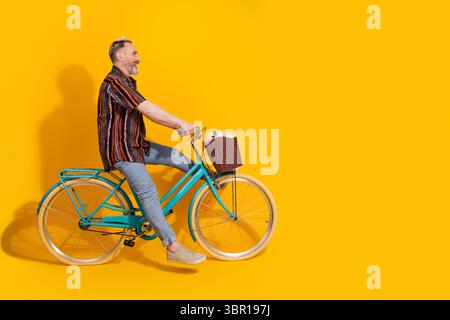 Charismatischer Mann mittleren Alters, der auf einem Retro-Fahrrad vor leuchtendem gelbem Hintergrund mit stilvoller Kleidung und fröhlichem Auftreten fährt Stockfoto